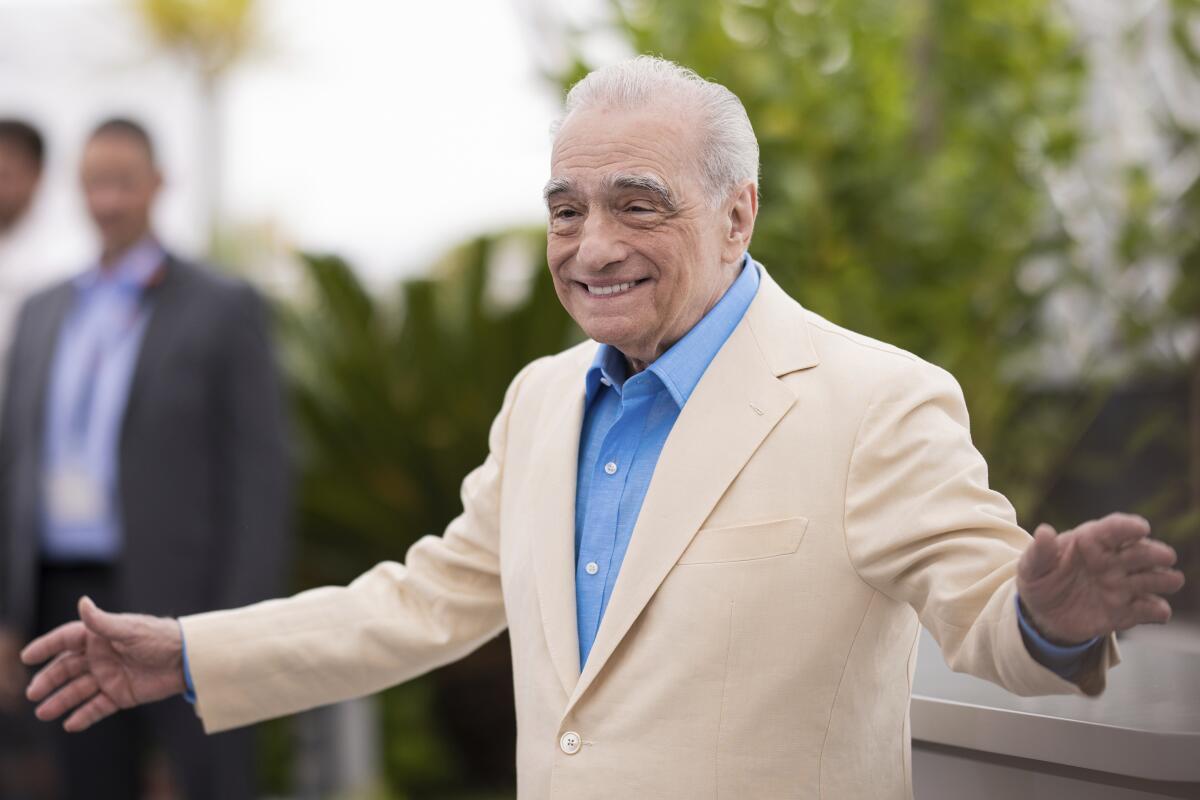 Martin Scorsese in a cream blazer and blue shirt, smiling with his arms raised at his sides