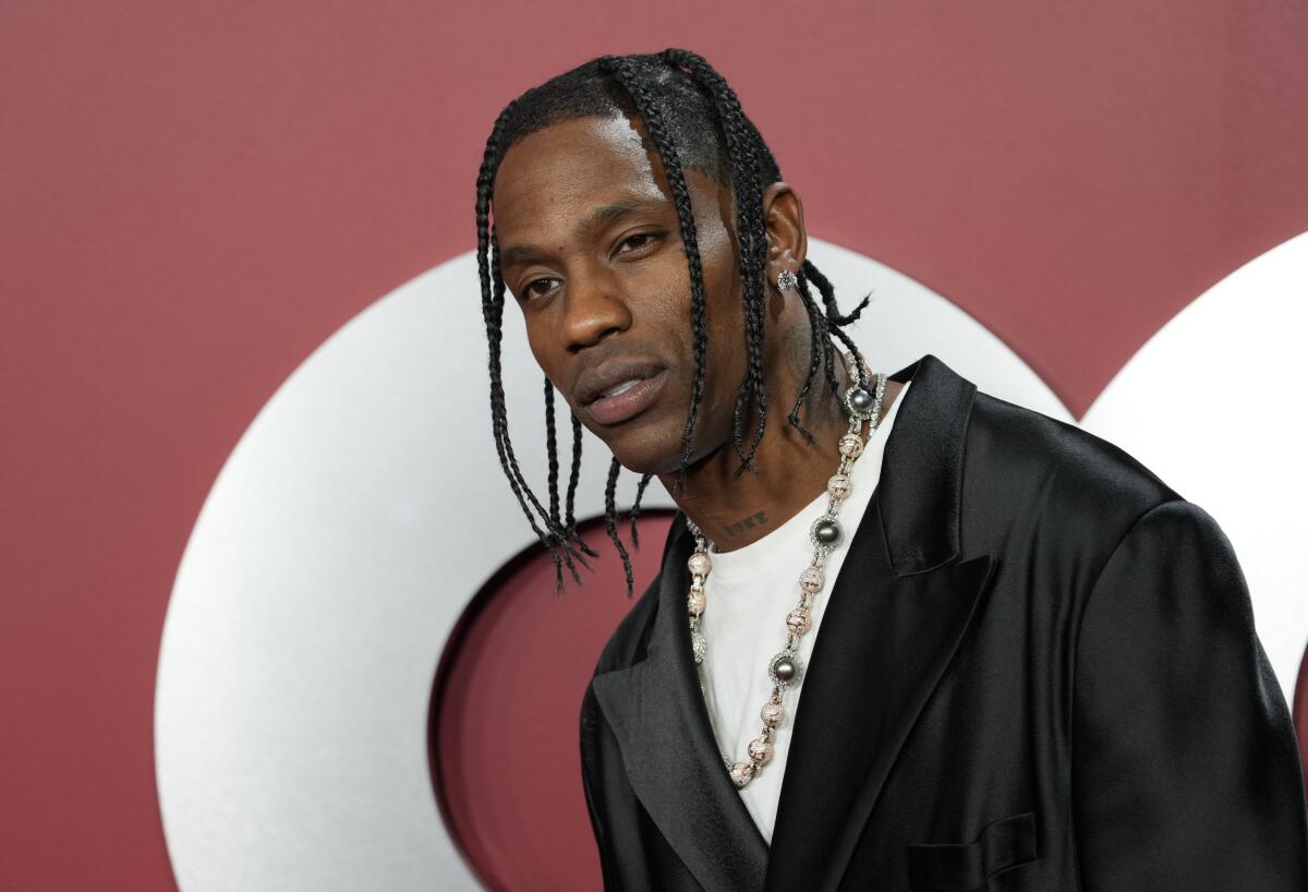 Travis Scott wears a white shirt and dark jacket with a necklace while standing in front of a red backdrop