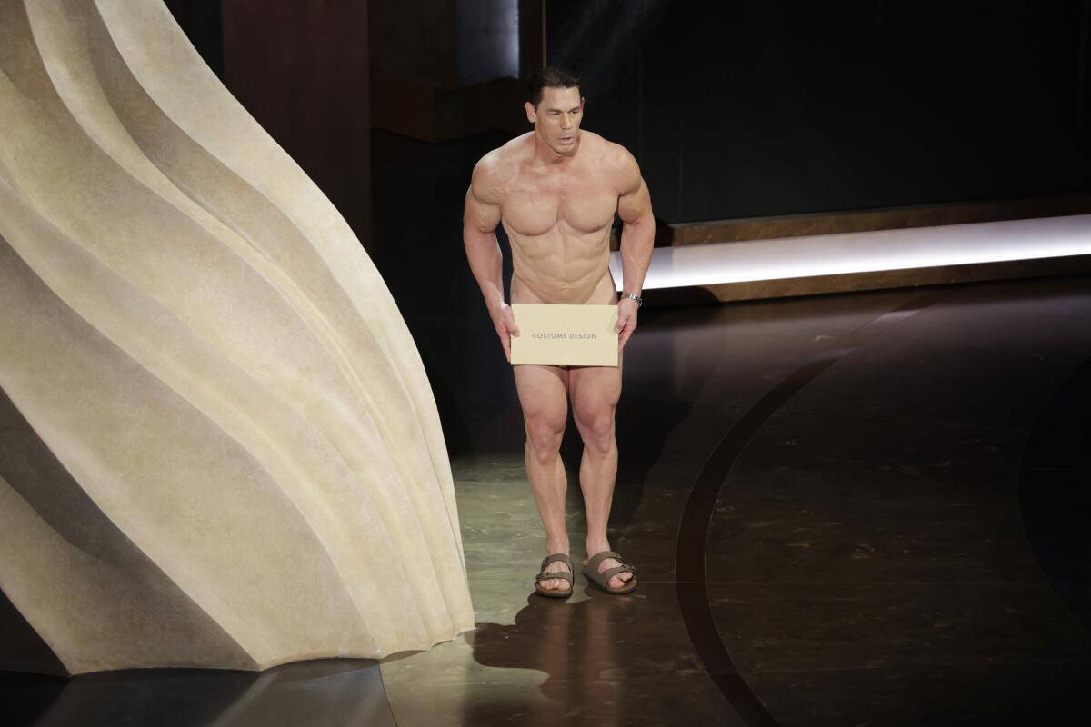 John Cena naked with an envelope over his groin and sandals standing on a stage next to a large beige setpiece
