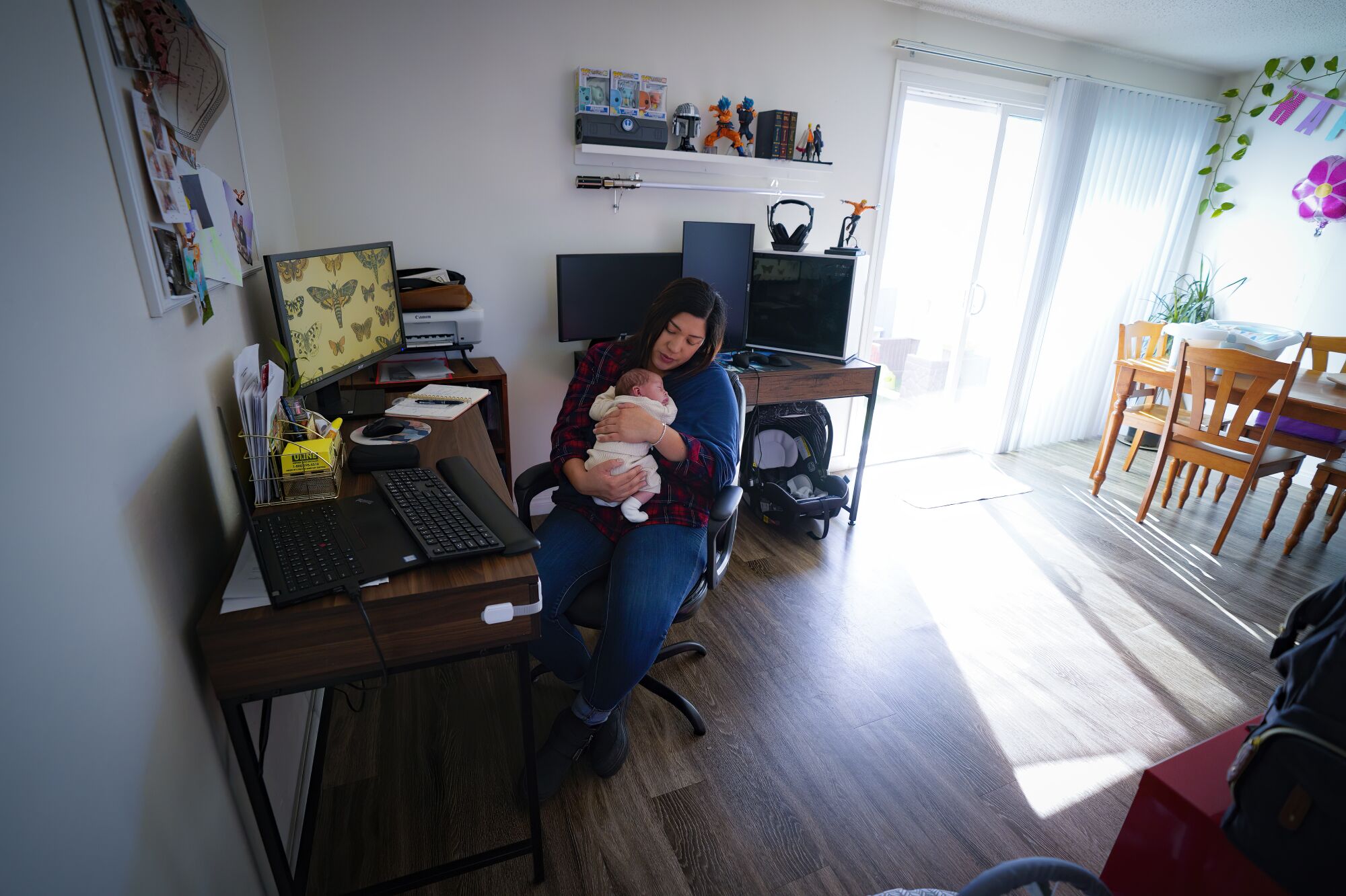 A woman sits at a desk in her living room, cradling a baby.