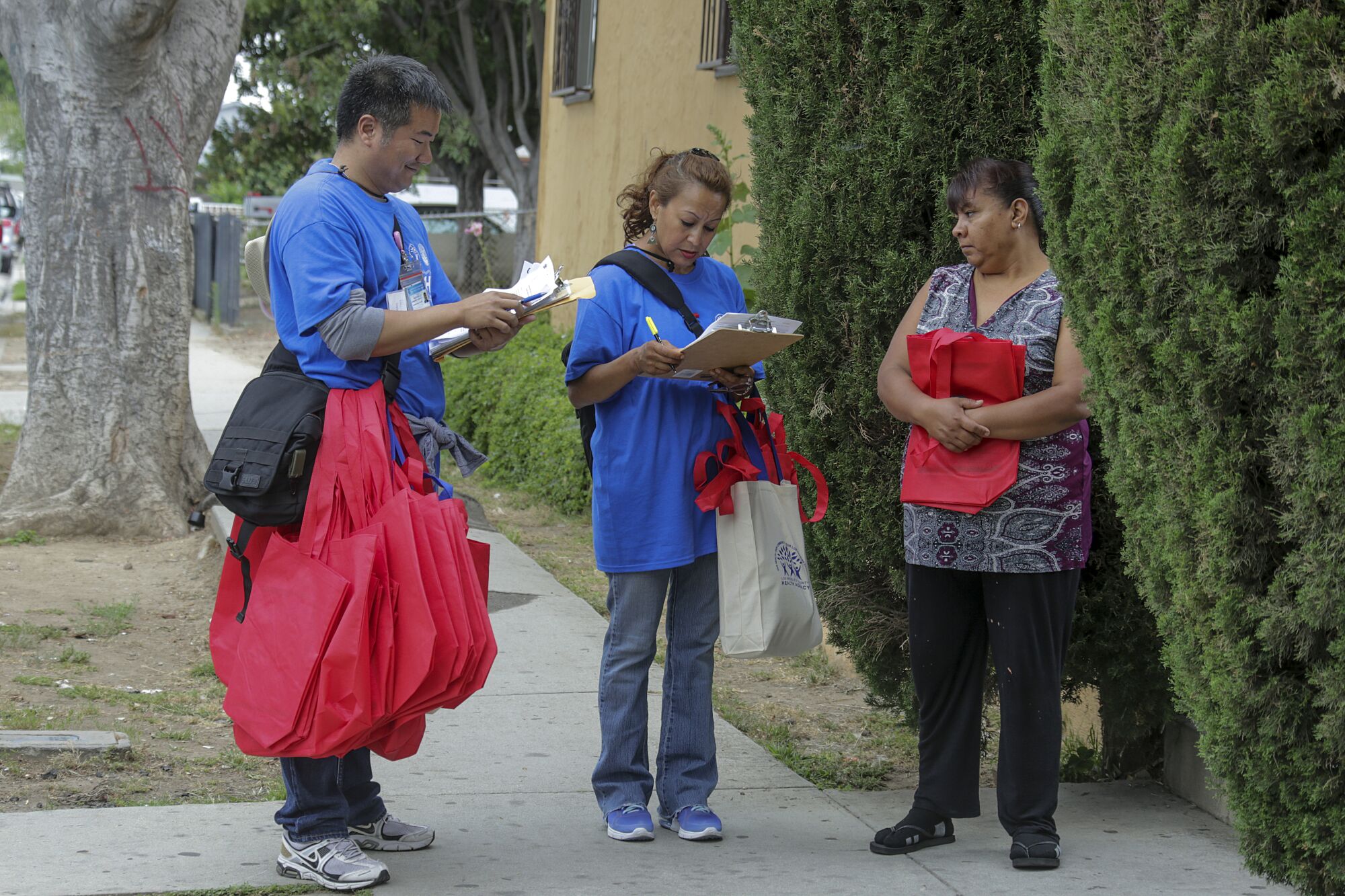 A man and woman carrying clipboards and red tote bags talk to a woman outside a residence.
