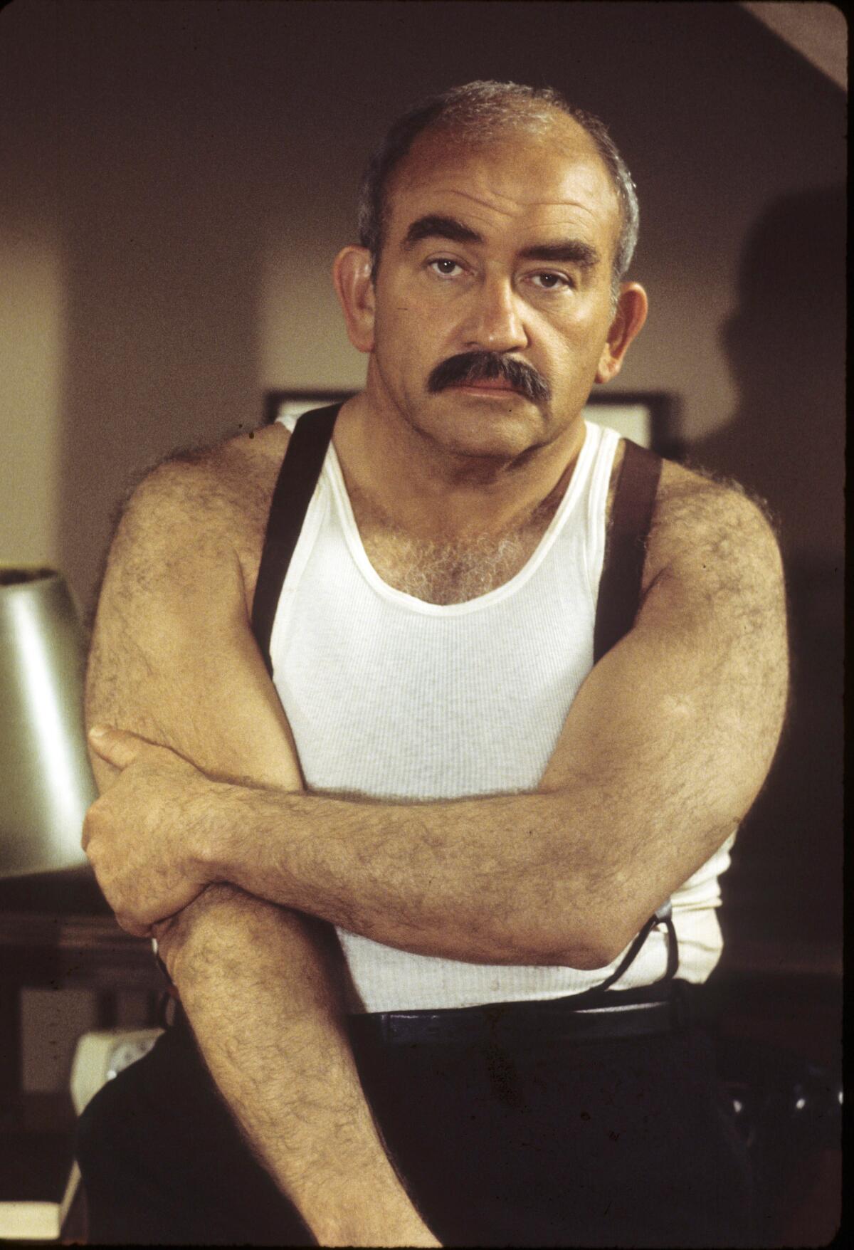 A man with a mustache wearing a white tank top and suspenders