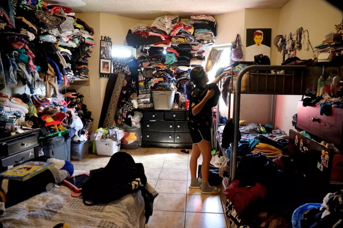 A young woman stands alongside a bunk bed in a room with clothing and belongings stacked to the ceiling on all sides.