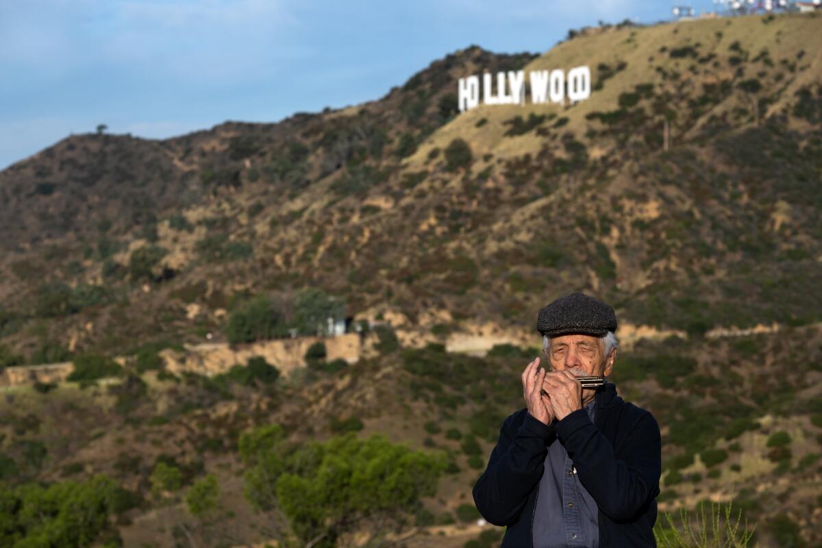 A man plays a harmonica in front of a hill with his "hollywood" sign.
