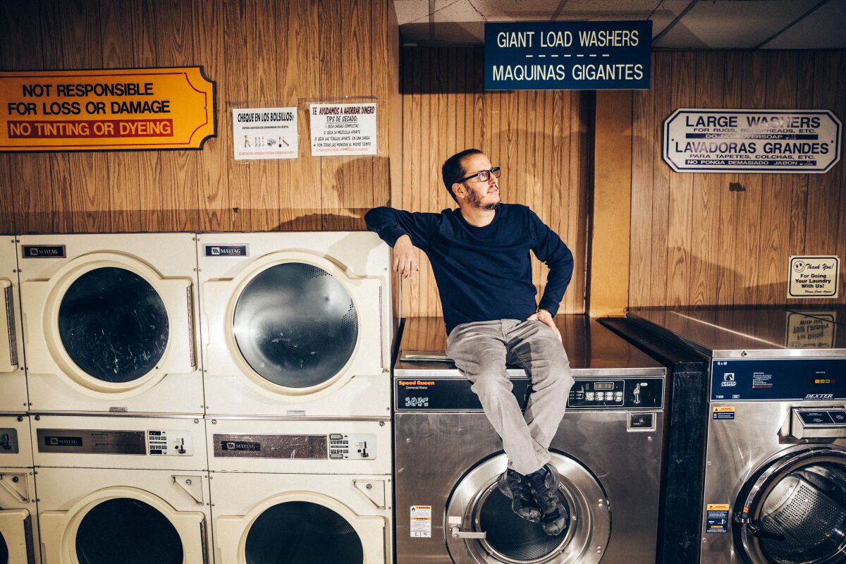 A man sits on a giant washer inside a laundromat.