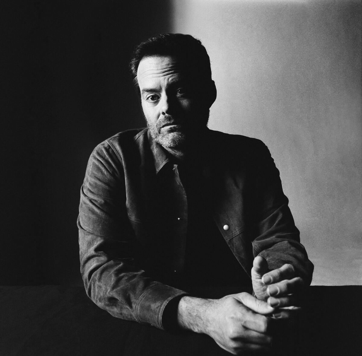 Bill Hader sits at a table for a black and white portrait. 