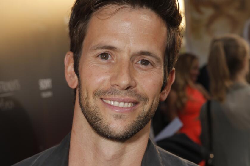 A man with short brown hair and stubble smiling and wearing a black leather jacket and a grey shirt. 