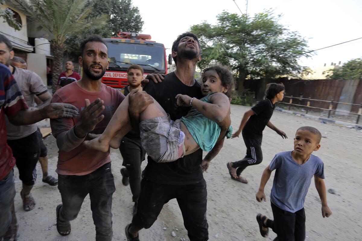 A young man carries a wounded child through a street while other people run nearby