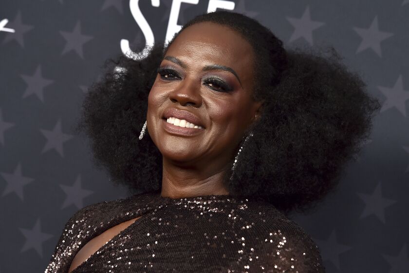A woman in a glittering formal gown and big hoop earrings smiles broadly upon arrival at an awards show