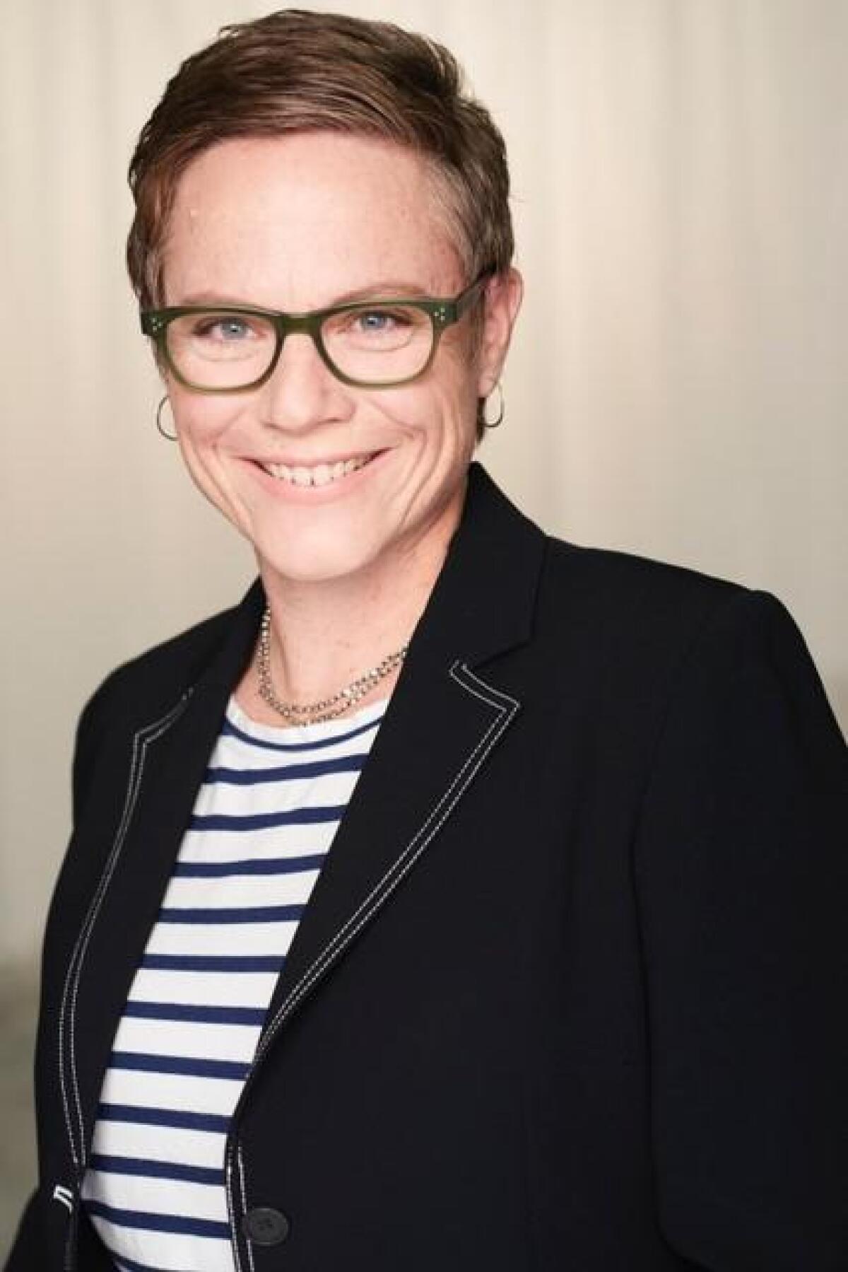 A person with short hair, glasses and earrings in a striped shirt and jacket