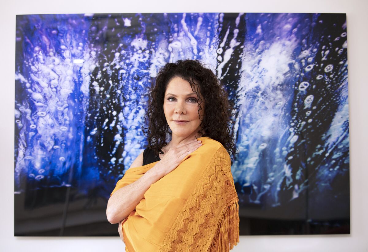 Author María Amparo Escandón wears a gold shawl and stands in front of a blue painting.