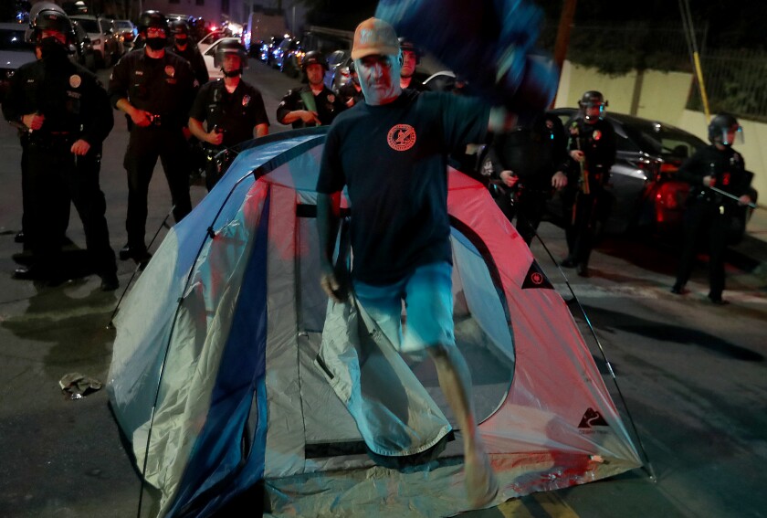 A man steps out of a small tent on a road in front of a line of police in riot gear.