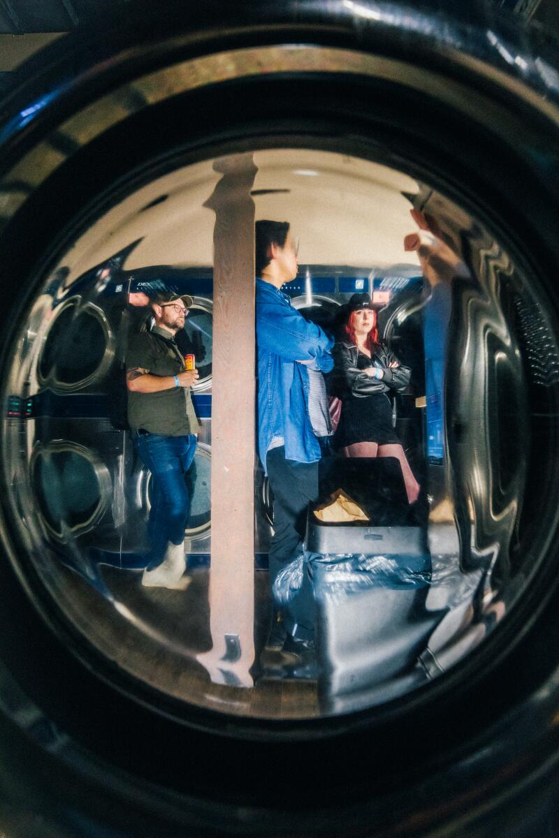 People stand in a laundromat in a view through a round, portal-like laundry machine door.