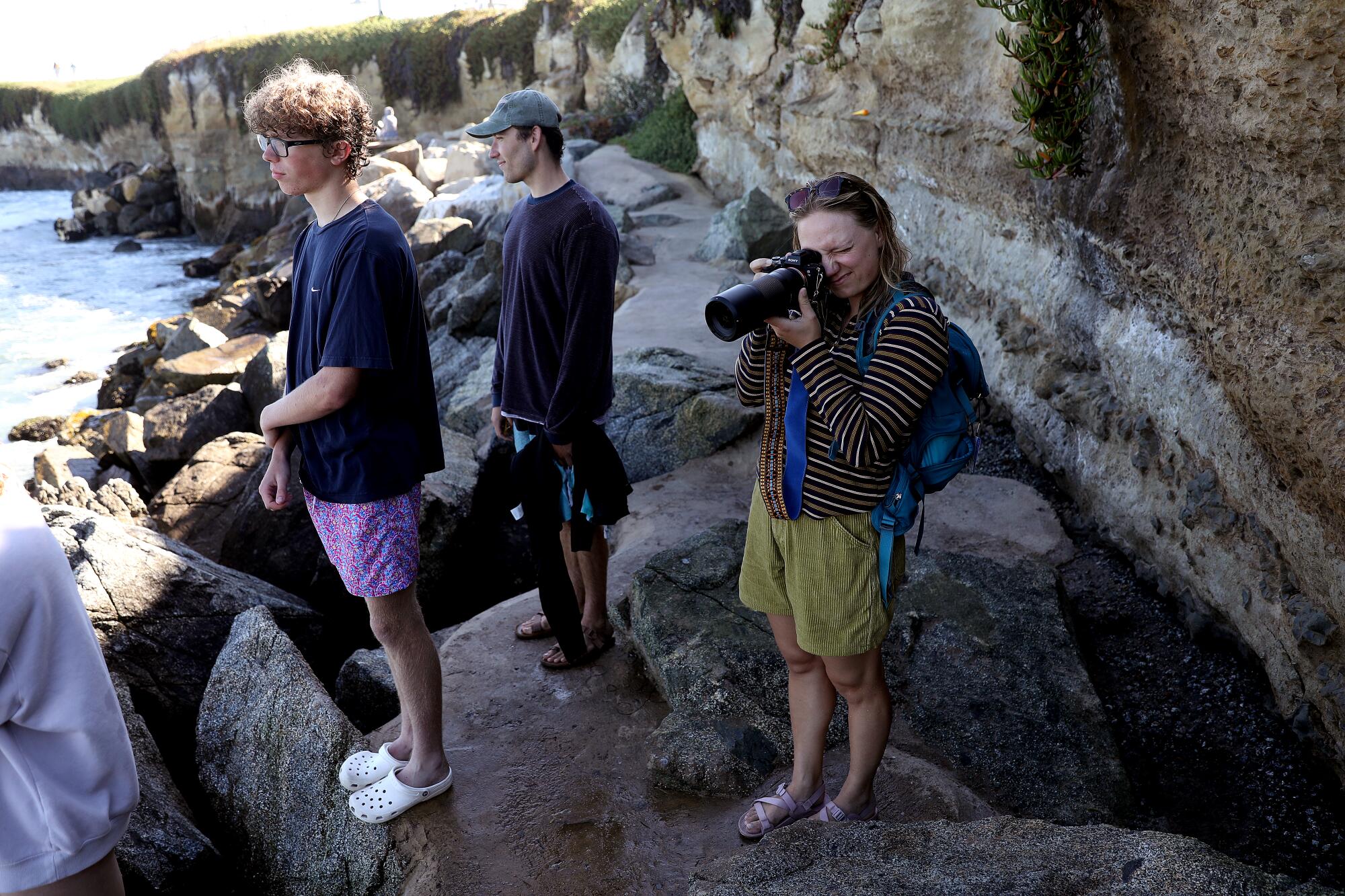 A group of people stand on rocks by the ocean and look out to sea. One of them holds a camera to his eyes.