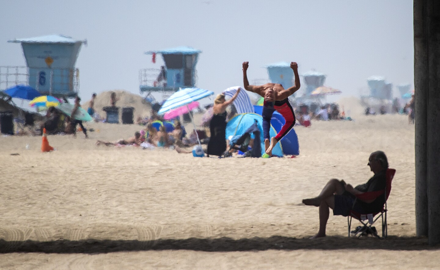 HUNTINGTON BEACH, CA -- THURSDAY, APRIL 30, 2020: Steve Christopher, right, reads a book in the shade of the pier as a beach-goer does flips across the beach on the last day of open beaches in Huntington Beach after Gov. Newsom announced the "hard close" of all Orange County state and local beaches Thursday, April 30, 2020. "I think it's what needs to be done," Christopher said. "I saw it last week and it was packed." Newsom's retaliation came after thousands of Californians flocked to the shoreline there over the weekend in defiance of a statewide stay-at-home order enacted to stem the spread of the coronavirus. "When you contact the police chiefs to enforce your will as a leader-in my book you establish a police state" said Huntington Beach resident Pete Hamborg. "It's a moment that Gavin Newsom is taking away from us and are the very moments that empower us to deal with this crisis on a day-to-day basis." "I think the beach, which belongs to all Californians, is critical to our emotional health and well-being." (Allen J. Schaben / Los Angeles Times)