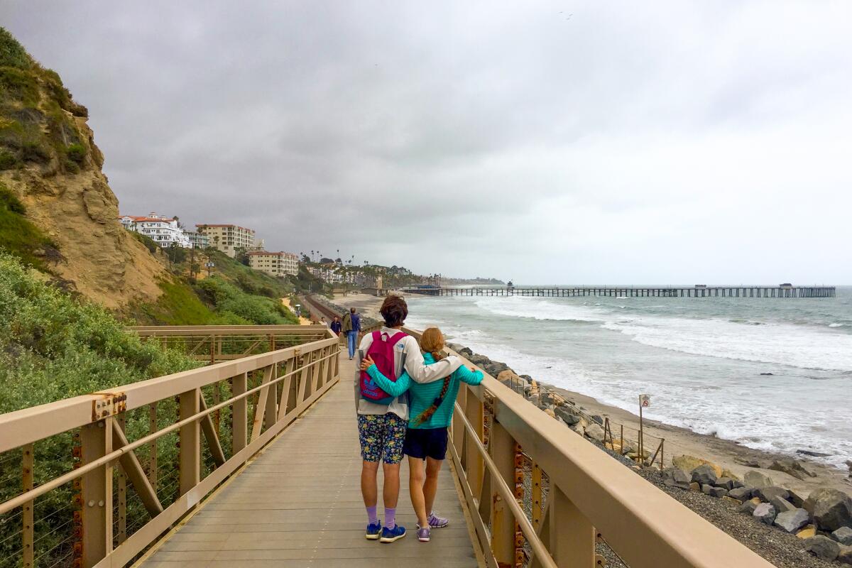 A couple hug while standing on a wooden walkway facing the beach 