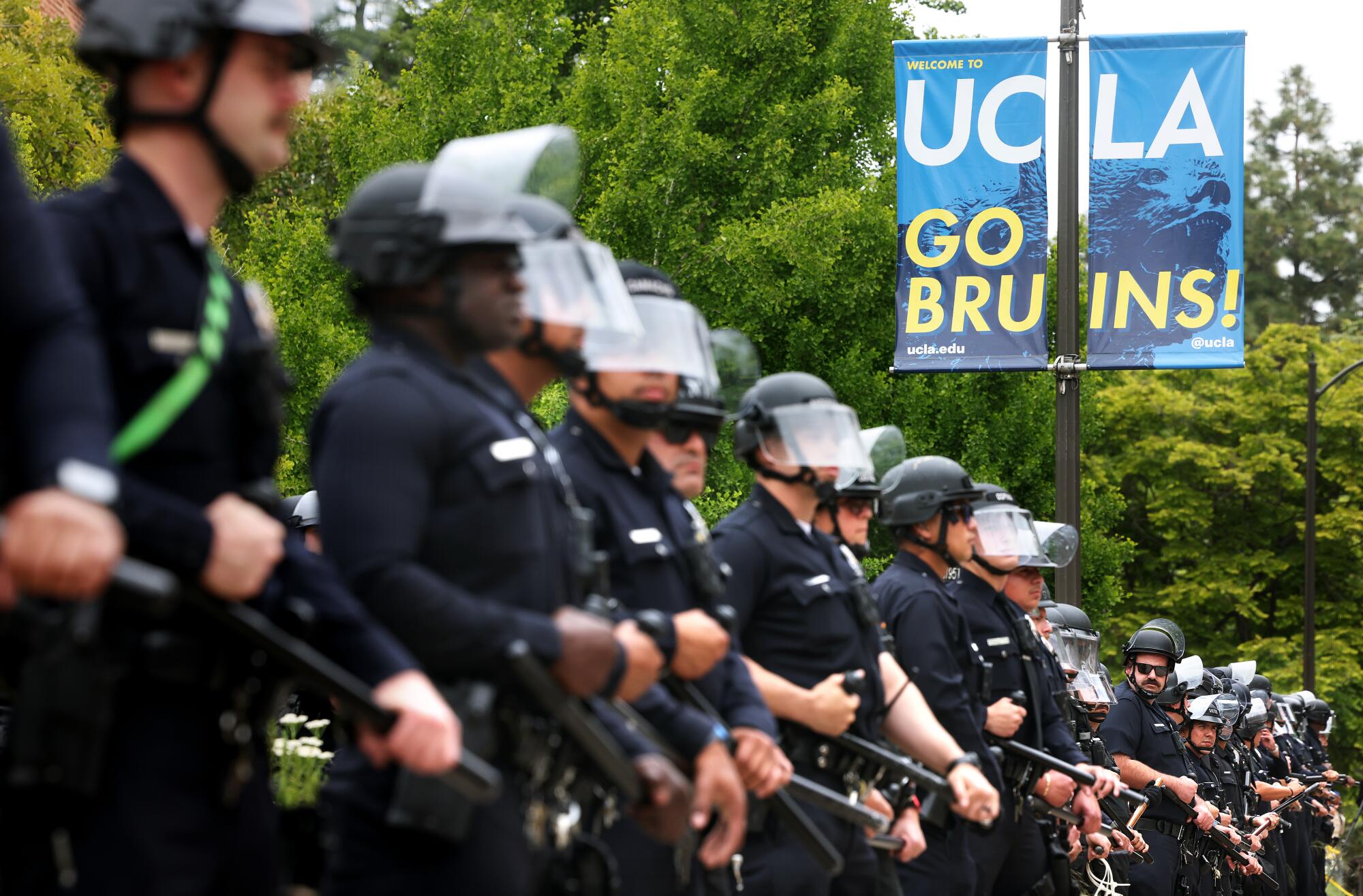 Officers in helmets and holding batons stand in a line near a sign that says "UCLA, Go Bruins!"