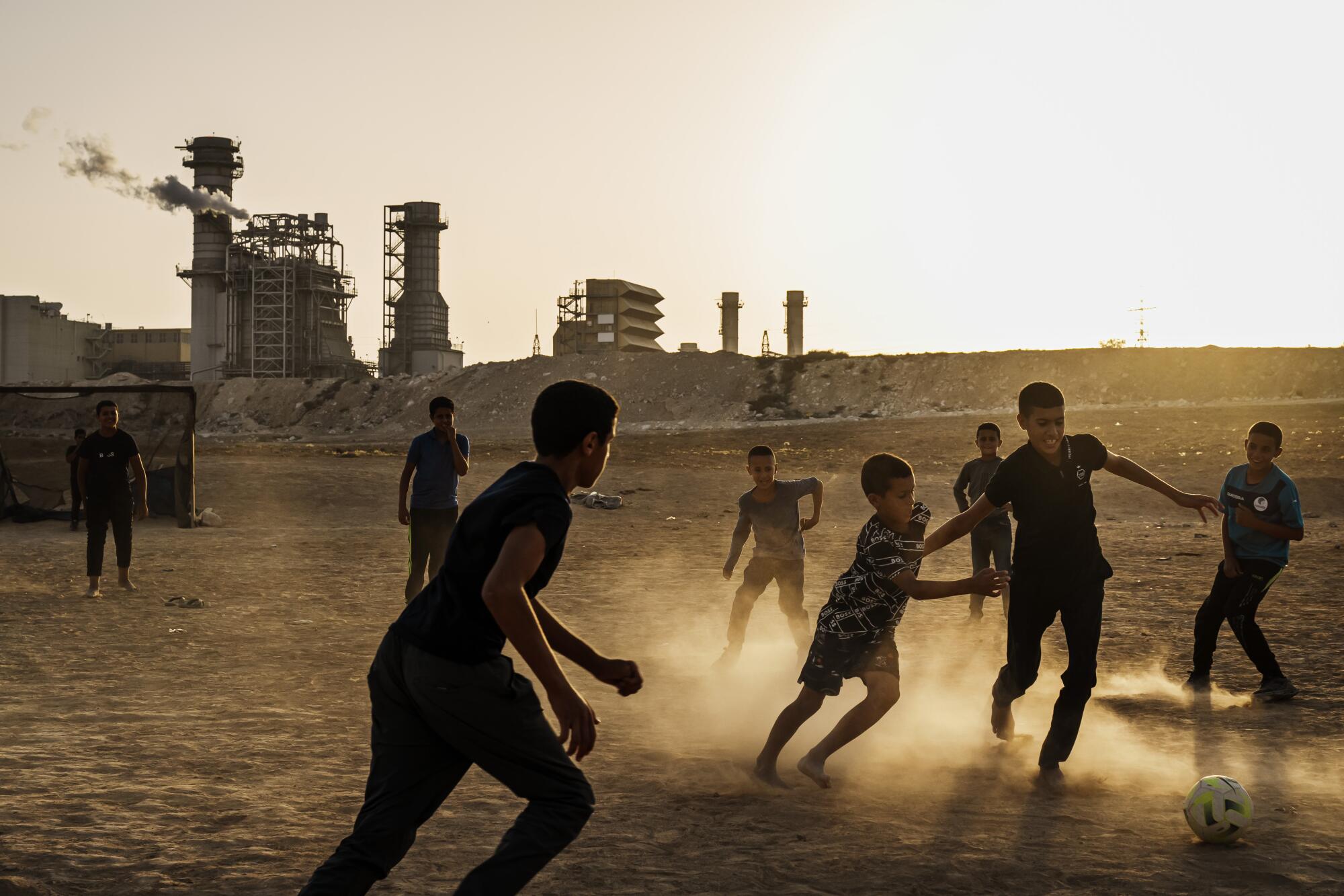 Children kick up dust as they play soccer at sunset in the desert, with a power plant in the distance.