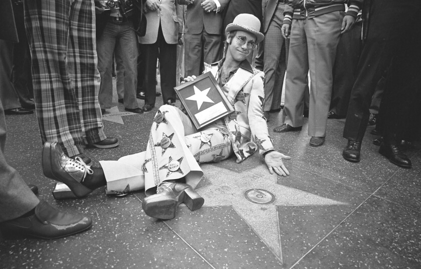 A man in platform shoes and a suit covered with stars and buttons with celebrity names poses on a sidewalk for a photo.