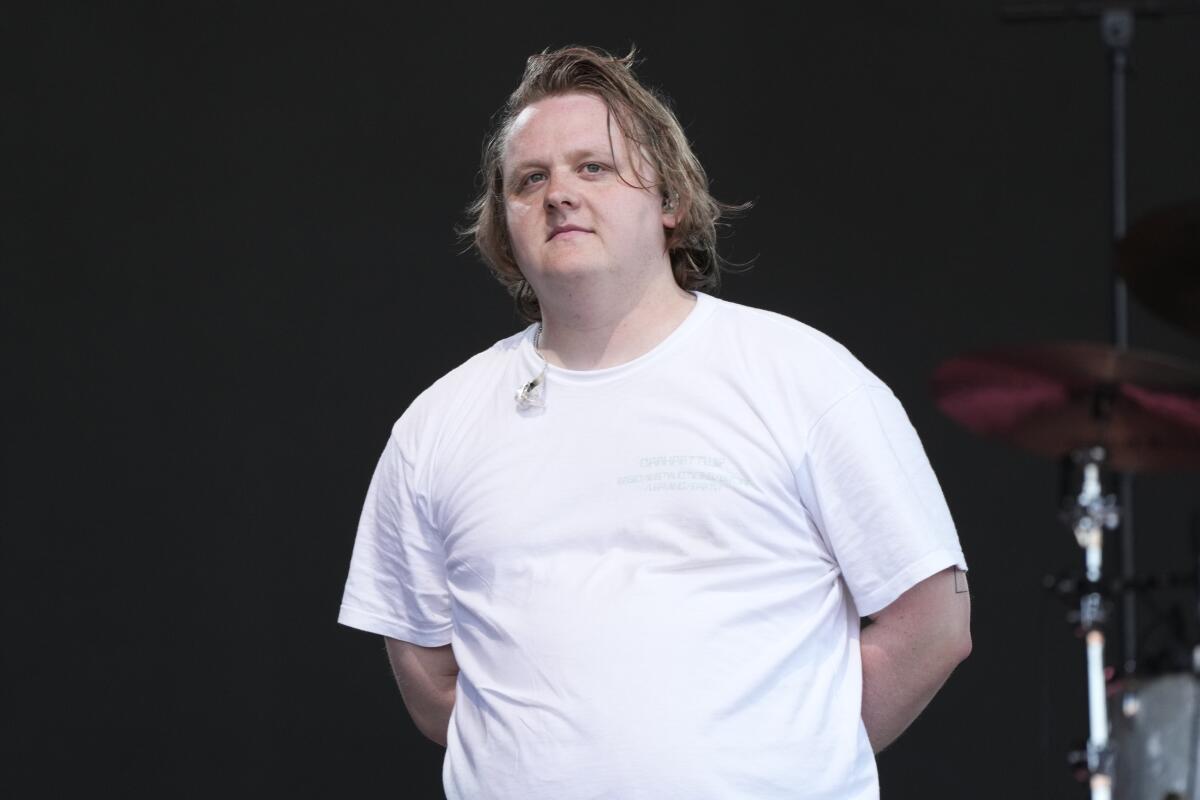 Lewis Capaldi wearing a white T-shirt standing on a stage with his hands behind his back 