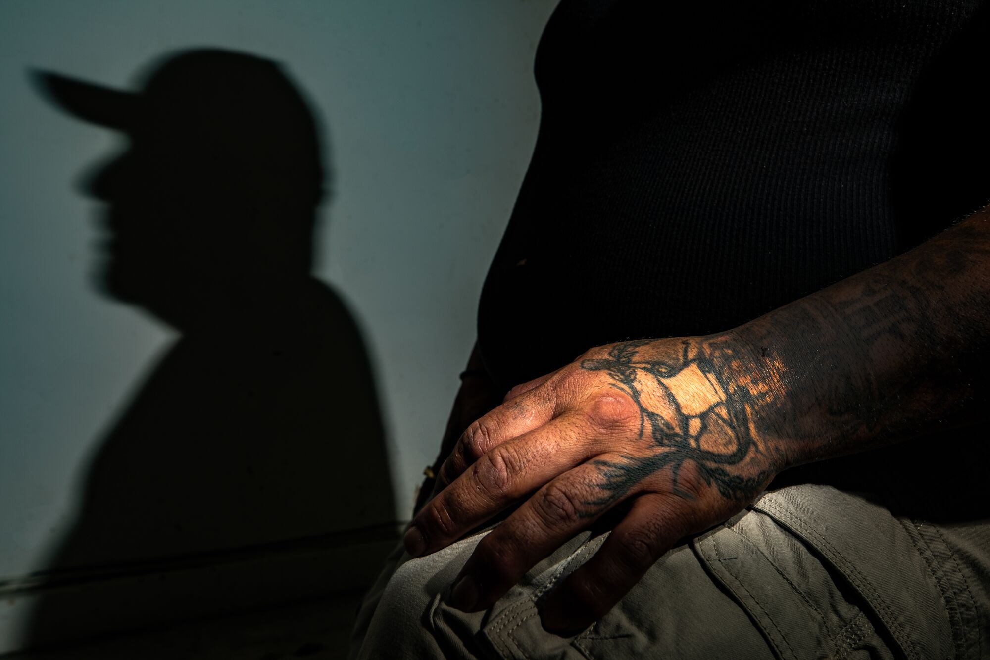 A close up frame of a tattooed hand resting on  a person's knee while his shadow is projected on the wall behind.
