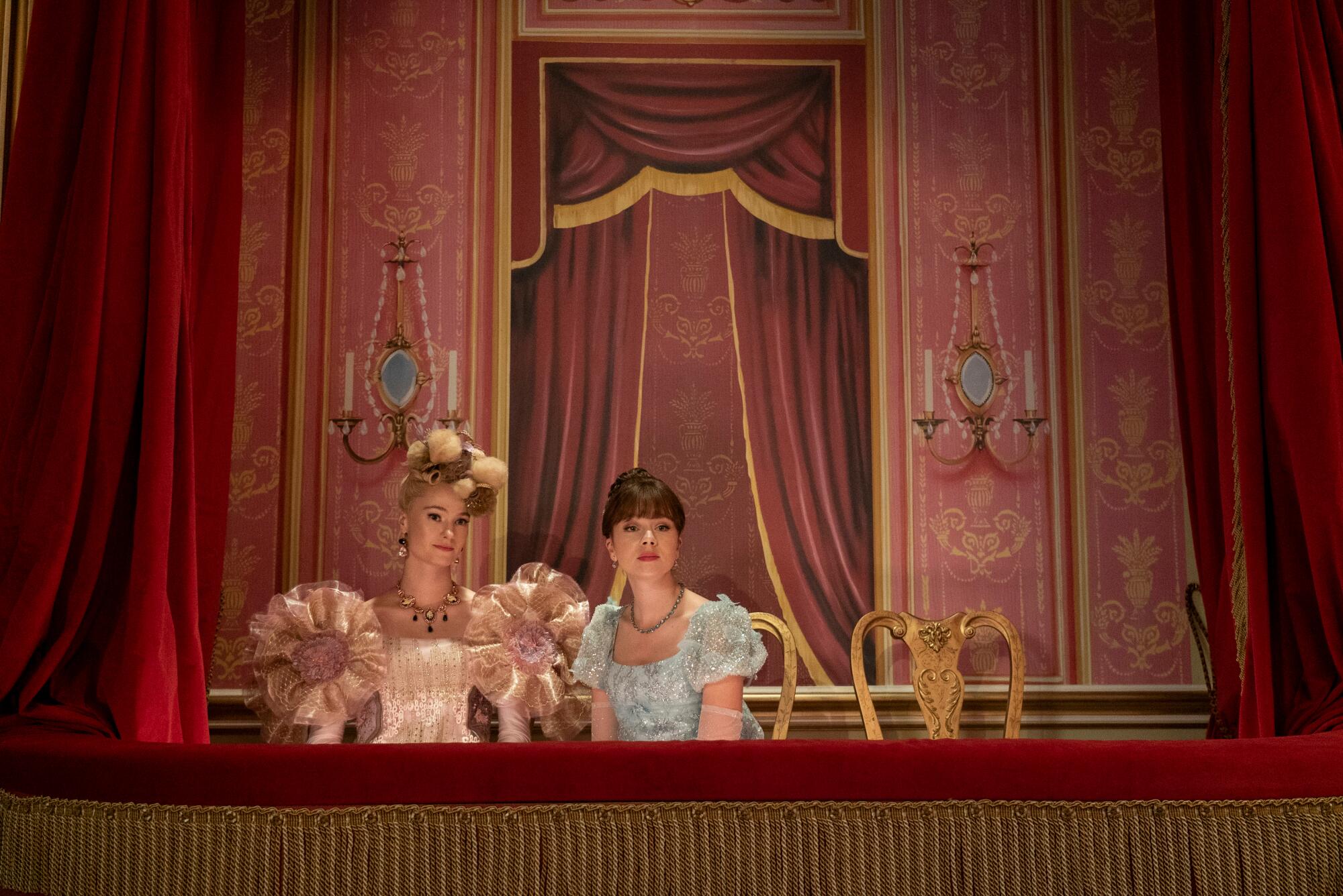 Two young women, wearing elaborate Regency-era dresses, sit in the balcony of a theater