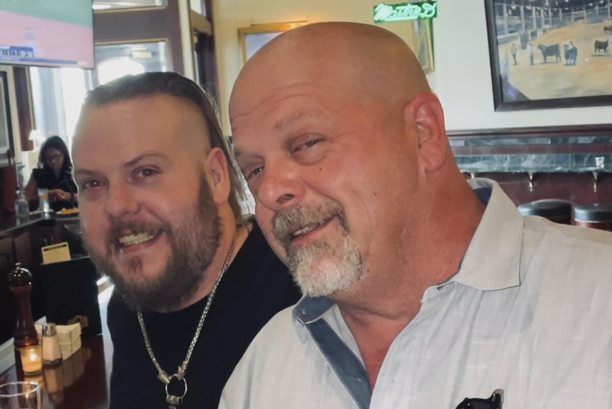 A bald man with a goatee in a white polo posing with a younger man in a black shirt and facial hair to his left