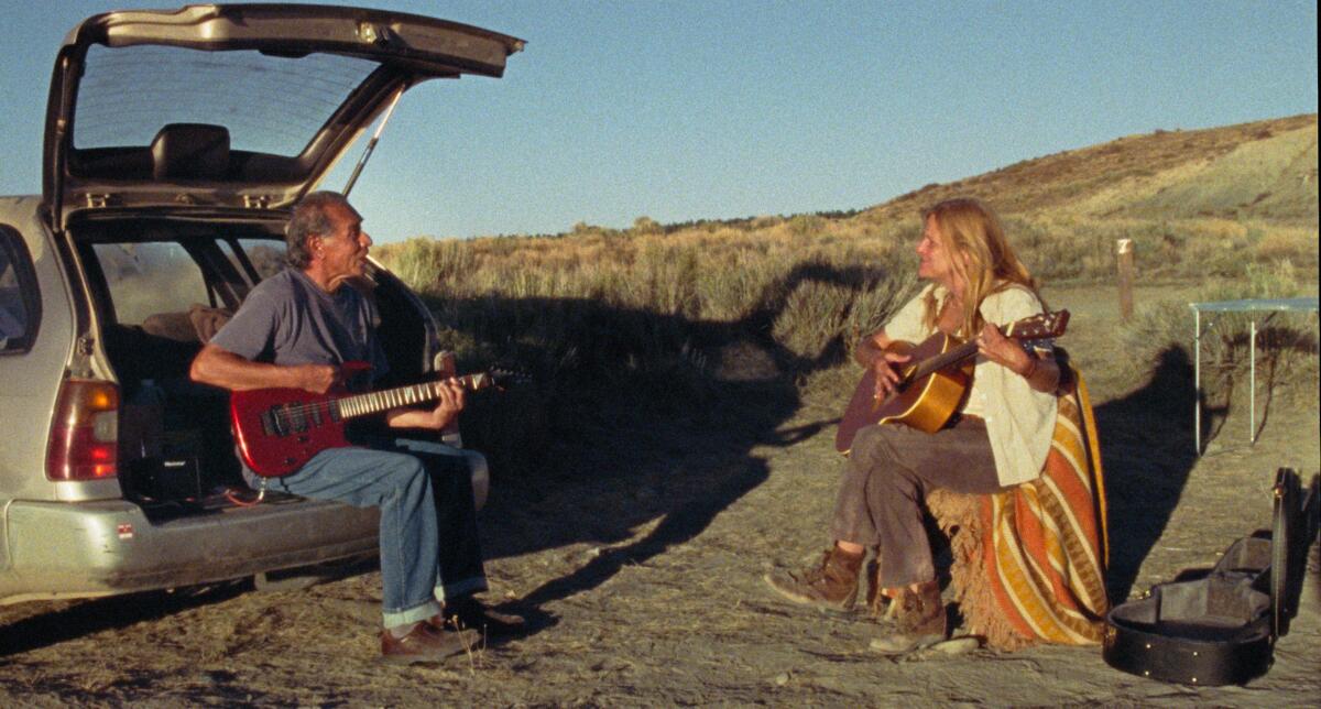 A man and a woman play guitar outdoors near a field. He sits in the rear of an open hatchback car.