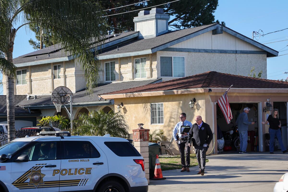 A police patrol car sits outside a suburban home. Two men walk along a driveway.