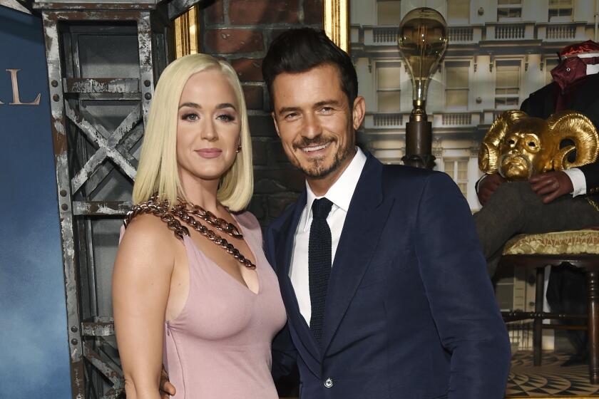 Katy Perry with blonde hair in a pastel pink dress posing next to Orlando Bloom with dark hair and blue suit