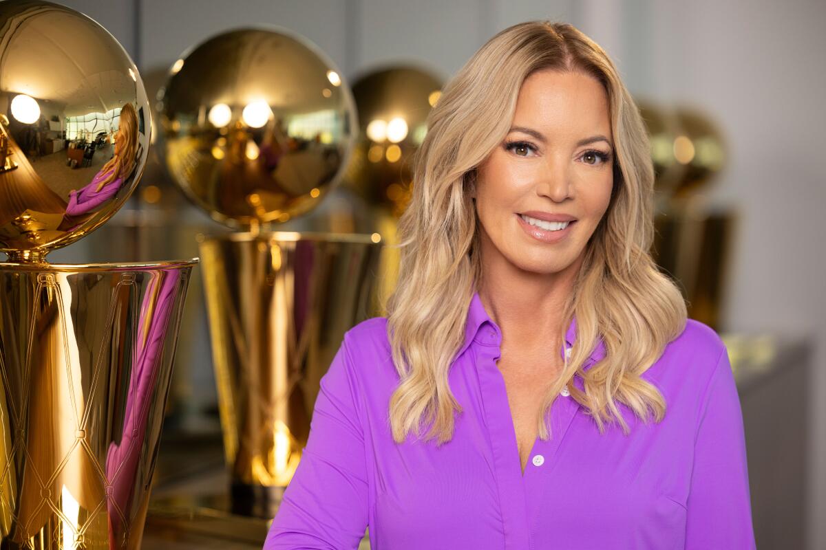 A smiling blond woman in a purple shirt stands in front of a row of golden trophies.