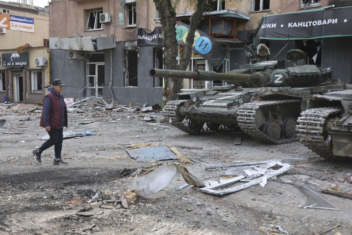 A woman walks by tanks in a destroyed city