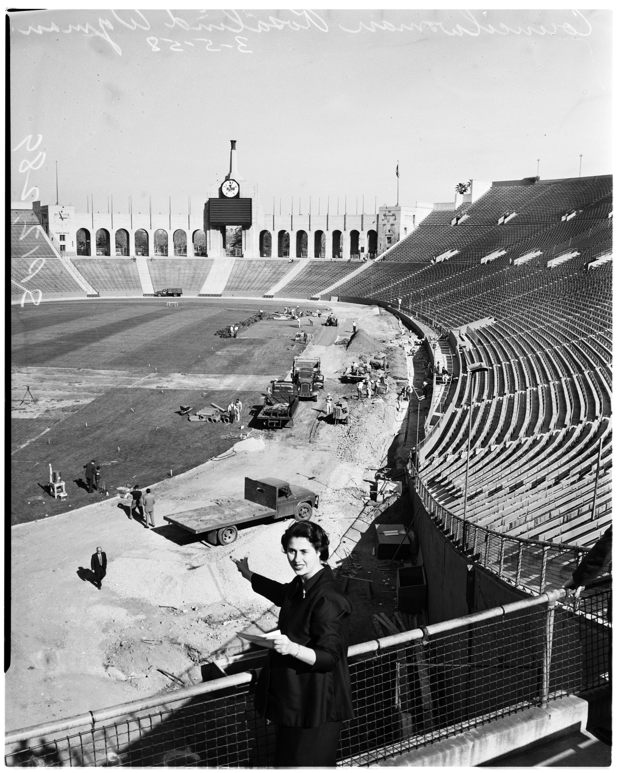 Rosalind Wyman in the stands of a coliseum while workers and vehicles were below 