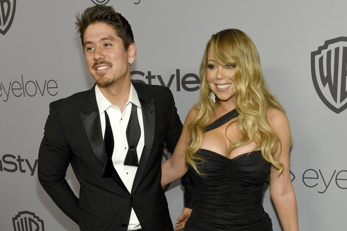 Bryan Tanaka, in a suit and undone bow tie, smiles while standing next to singer Mariah Carey, wearing a formal black gown