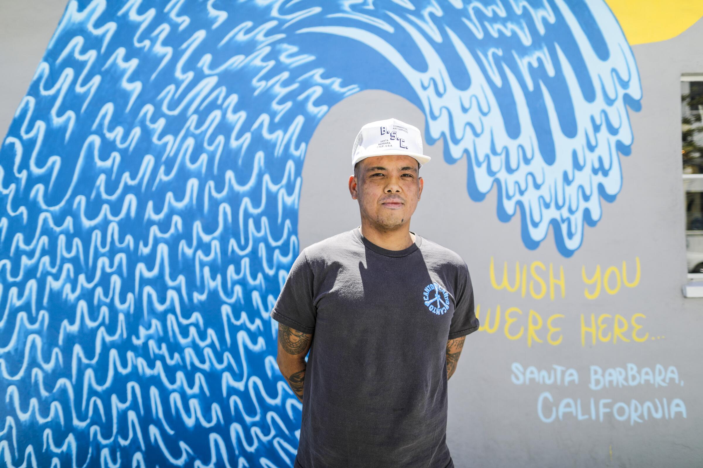 A young man in a T-shirt and baseball cap in front of a mural of a wave and the words "Wish you were here."