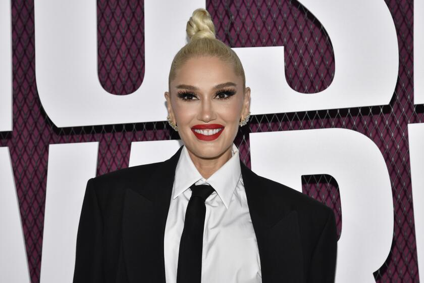 Gwen Stefani with her hair in a high bun and slicked back. She's wearing a black suit jacket, white shirt and black tie. 