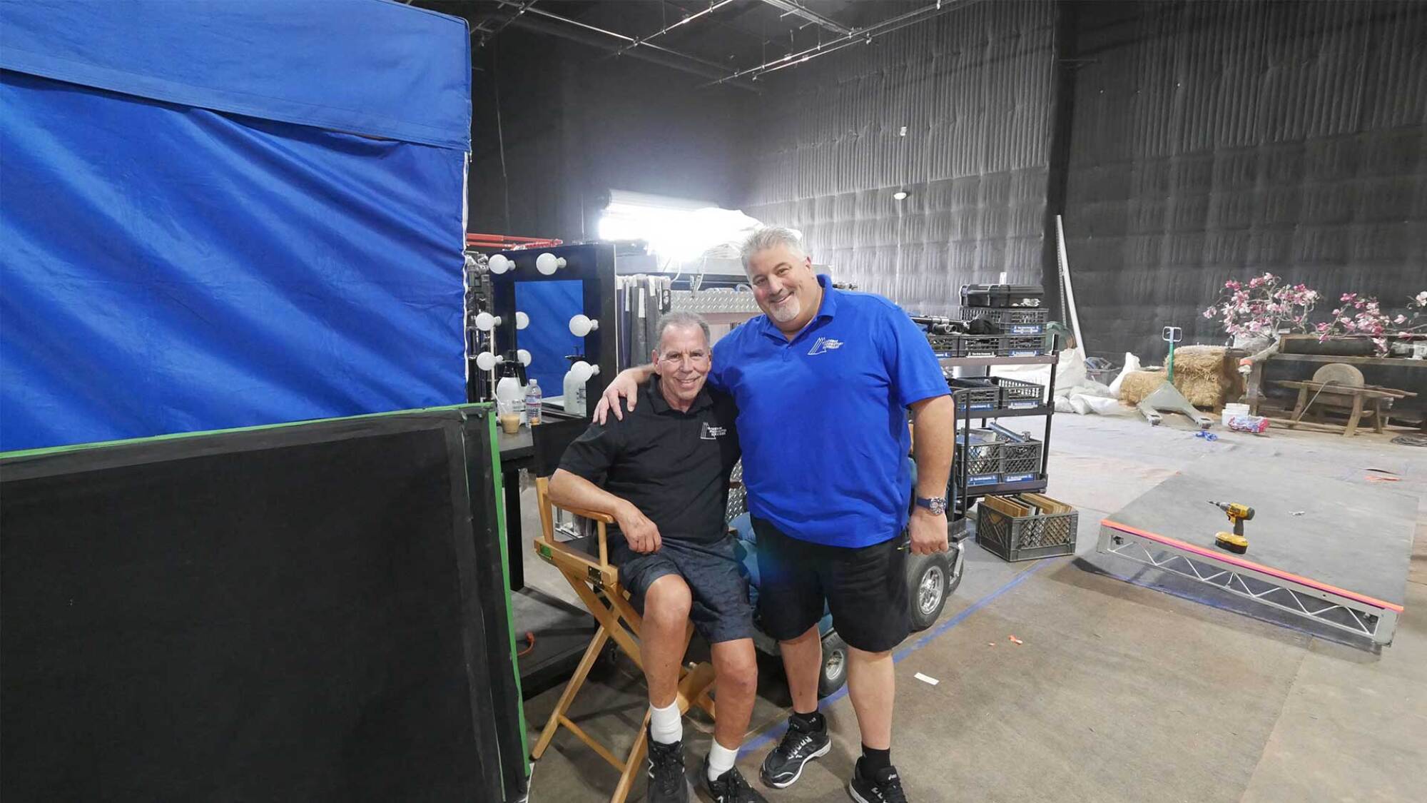 A standing man smiling and posing with his arm around a man sitting in a folding chair on a soundstage