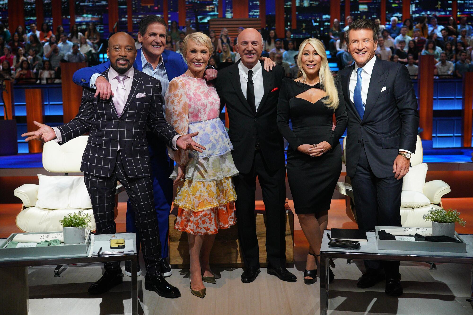 Six celebrity panelists in dress attire smiling for a group photo on set