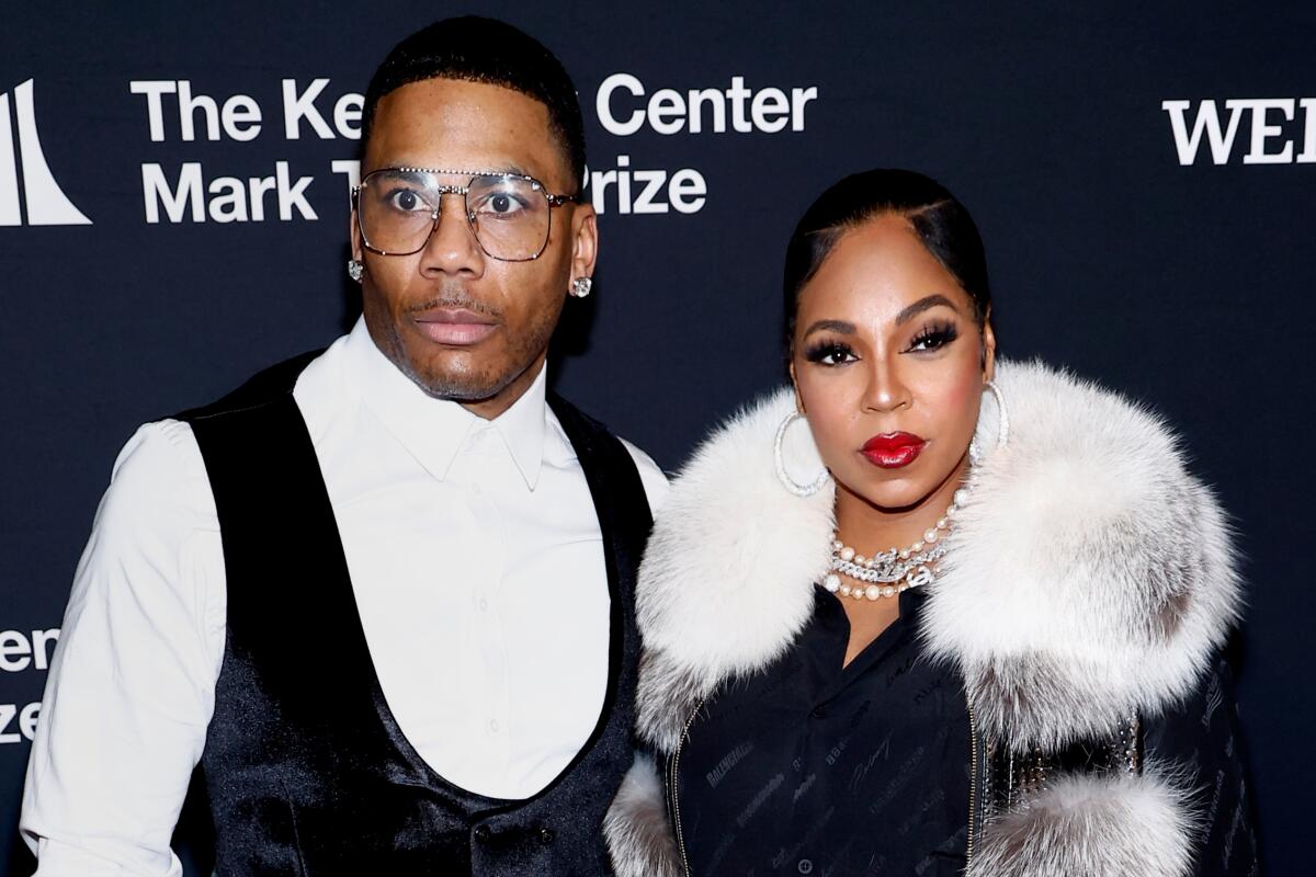 Nelly in a white dress shirt and a black vest standing next to Ashanti in a fur coat over a black dress
