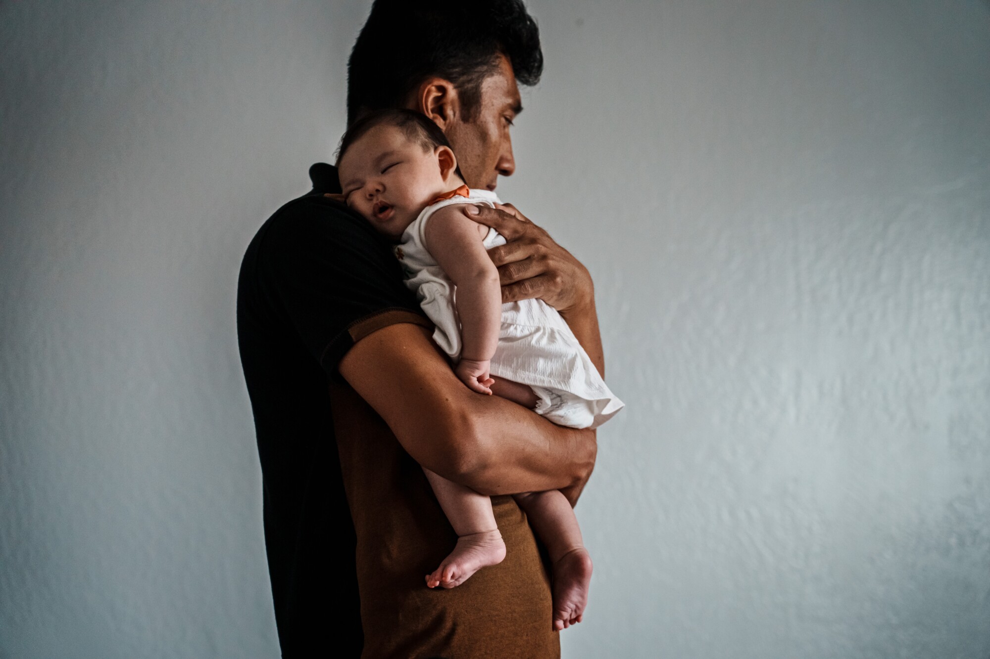 A dark-haired man holds a sleeping baby against his shoulder 