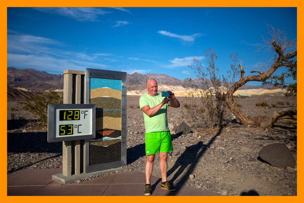 A man stands in a desert taking a selfie next to a digital thermometer that reads 128 degrees Fahrenheit