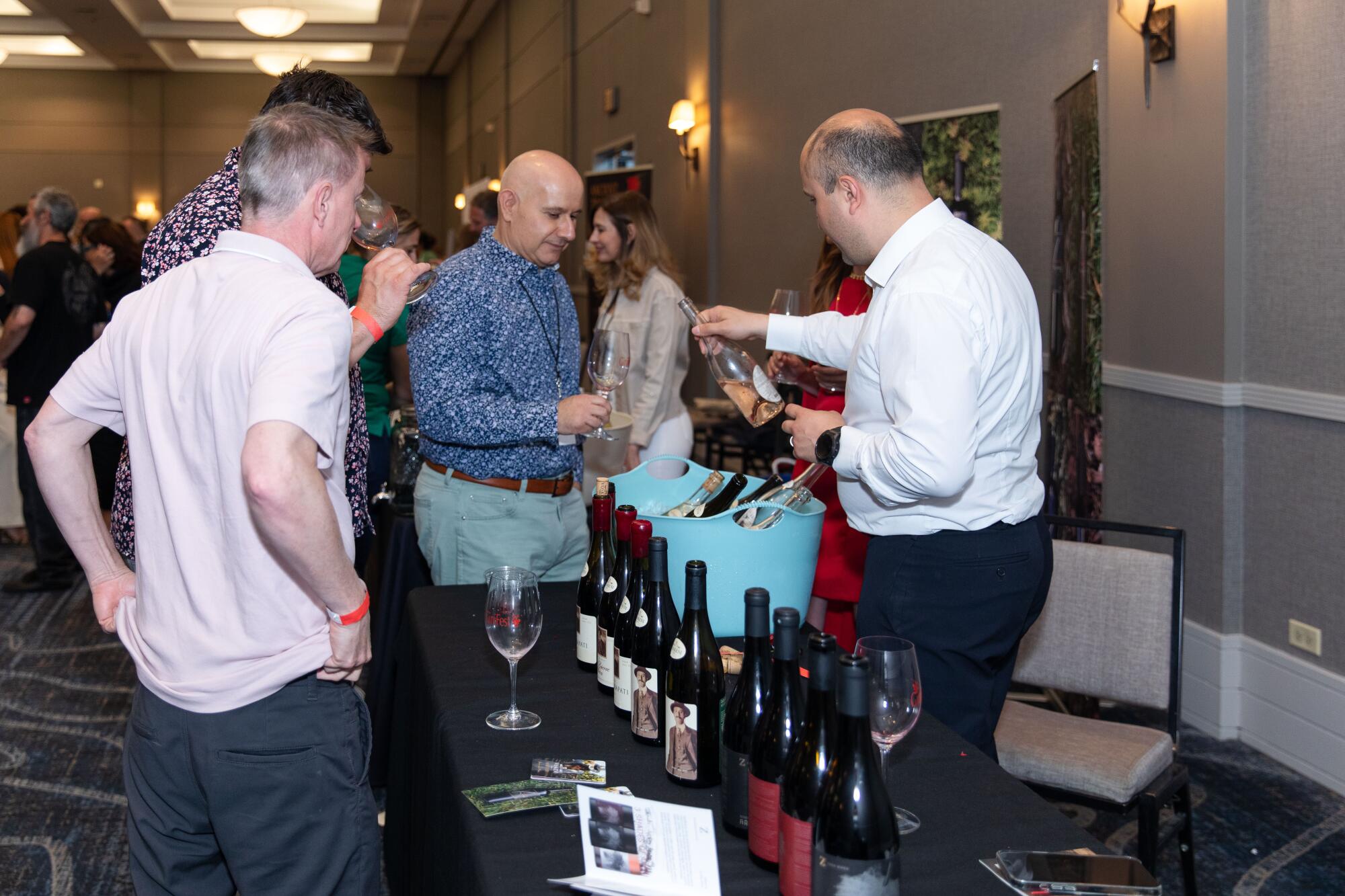 Several men stand examining wine bottles and tasting wine.