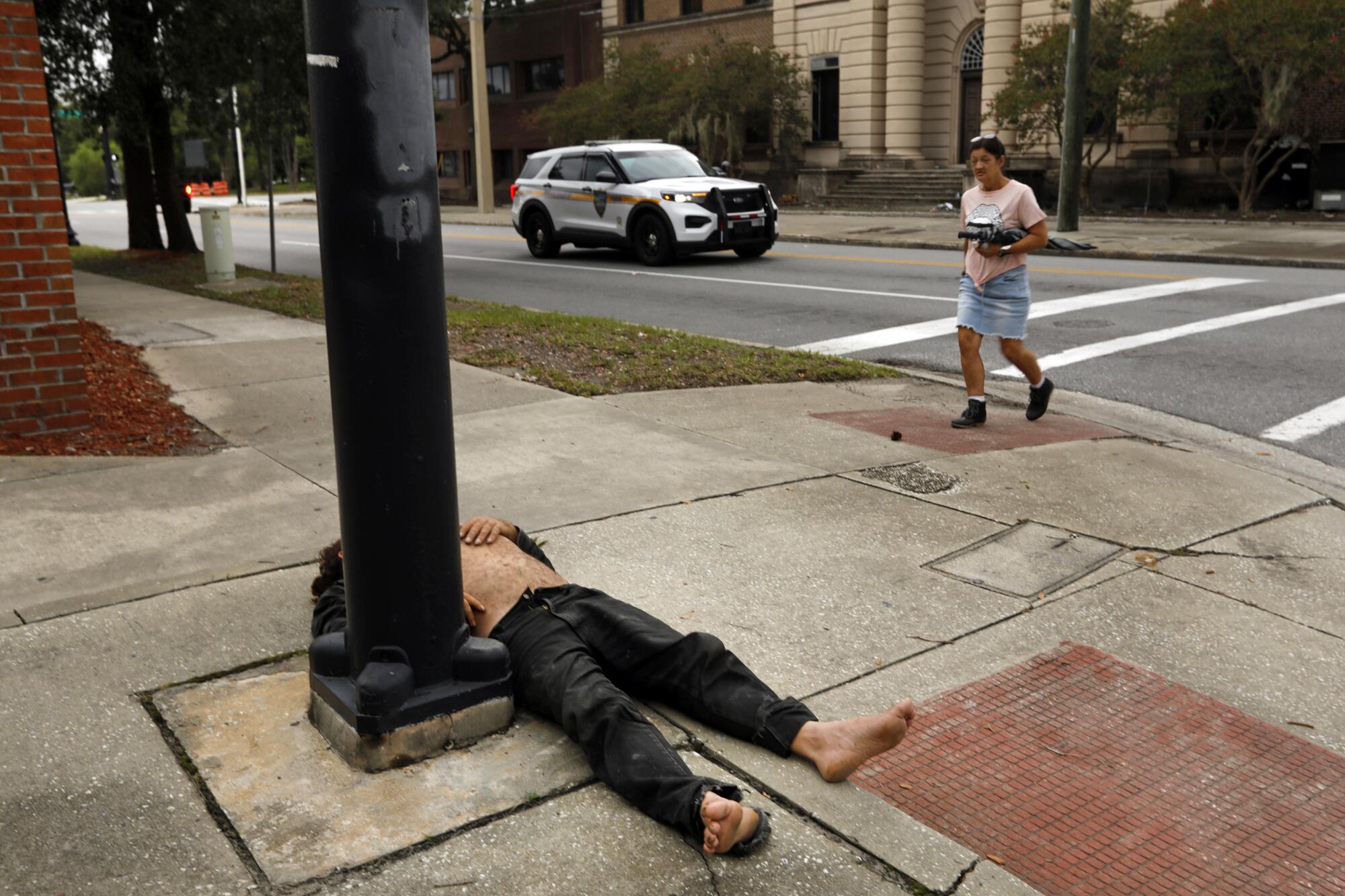 A man walks onto a street corner, near a shirtless person in dark pants lying next to a pole 