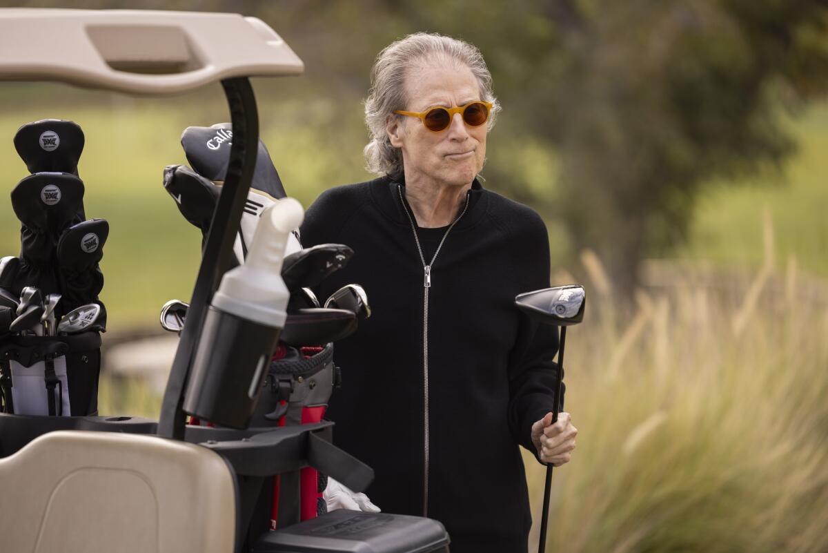 Richard Lewis, dressed all in black, holds a golf club and stands next to a golf cart. 