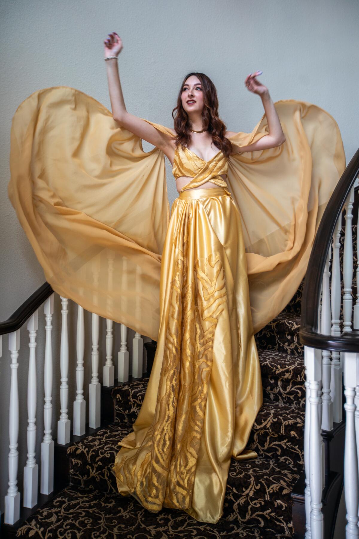 A woman on a staircase wears a gold, flowy, floor-length gown.