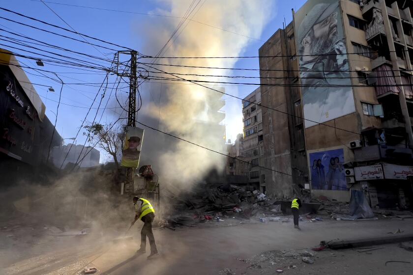 Workers clean a street under a giant portrait of the late Hezbollah military commander Imad Mughniyeh, as smoke rises from a destroyed building that was hit by an Israeli airstrike in Dahiyeh, in the southern suburb of Beirut, Lebanon, Sunday, Oct. 20, 2024. (AP Photo/Hussein Malla)