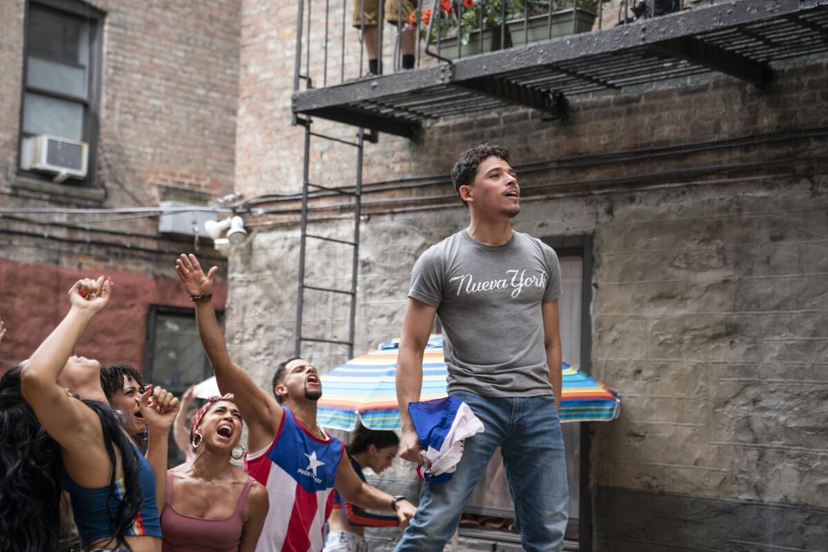 Anthony Ramos dances on a table in an alley wearing a grey T-shirt that reads "Nueva York"