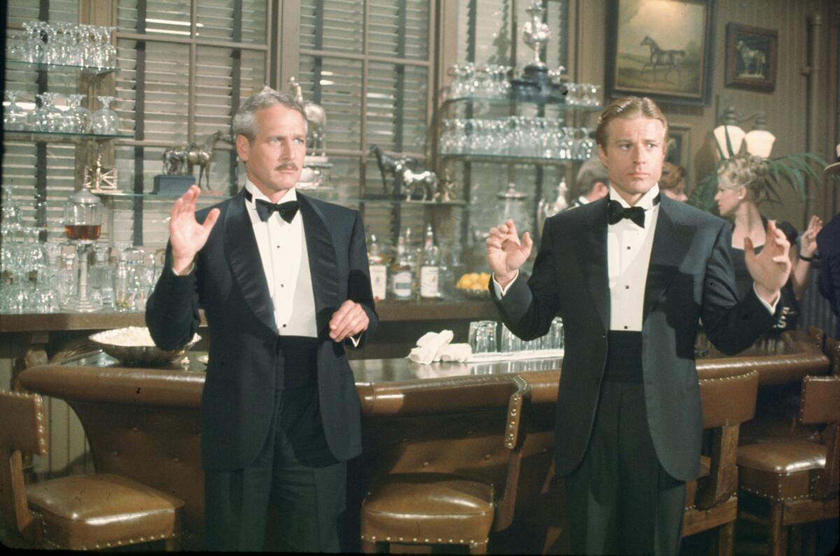 Two men in tuxedos stand next to a bar with their hands up.