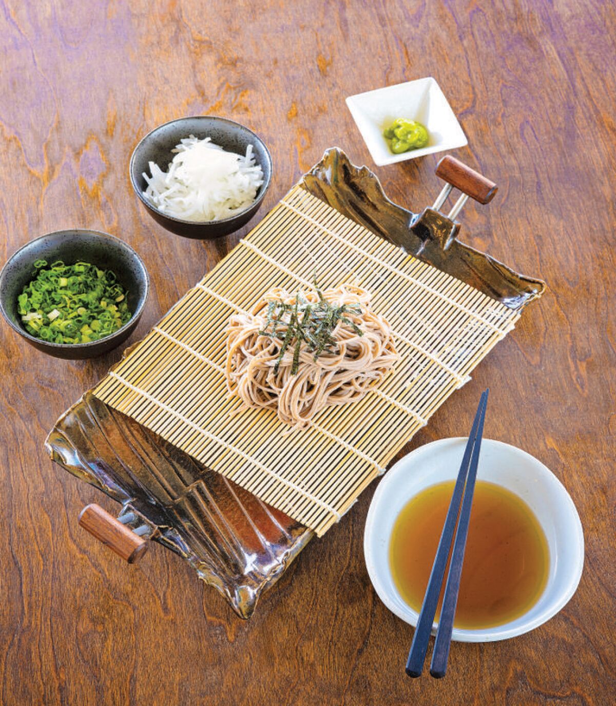 Cold soba noodles with dipping sauce and toppings are arranged on a table.