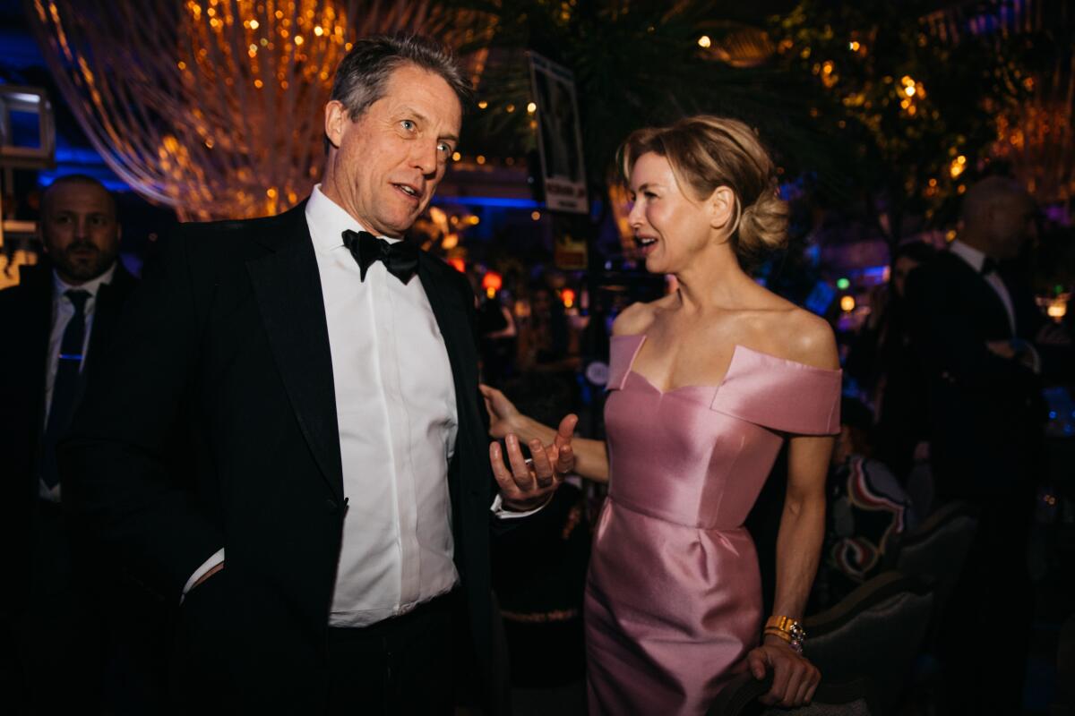 Hugh Grant in a tuxedo speaking with his arms out next to Renee Zellweger in a sleeveless pink gown
